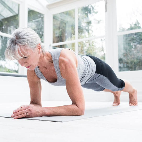 Frau in ihren 60ern, die zu Hause auf einer Yogamatte Pilates macht. Sie balanciert auf ihren Unterarmen und schaut auf den Boden.