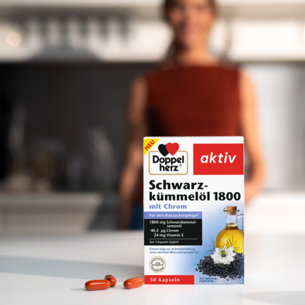 Das Produkt Doppelherz Schwarzkümmelöl 1800 auf einem Tisch, daneben liegen die Kapseln.