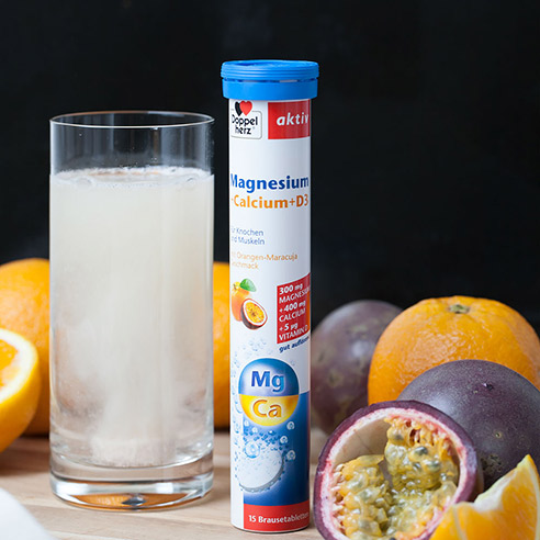 Doppelherz Magnesium + Calcium + D3 Brausetabletten, Brausetablette löst sich in Glas Wasser auf, daneben Orangen und Maracuja